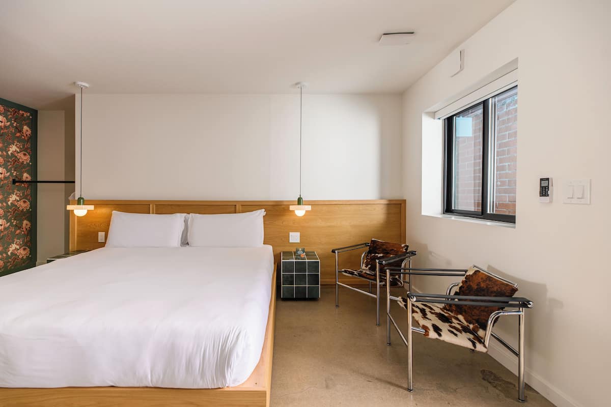 A hotel room with a large bed, a wooden headboard, a small table, a chair, and a window with a view of a brick building.