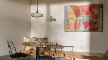 A dining area with a wooden table, two chairs, and a bench. There is a hanging light fixture and a colorful abstract painting on the wall.