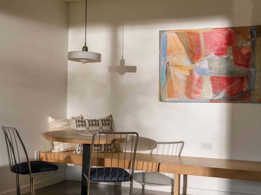 A dining area with a wooden table, two chairs, and a bench. There is a hanging light fixture and a colorful abstract painting on the wall.
