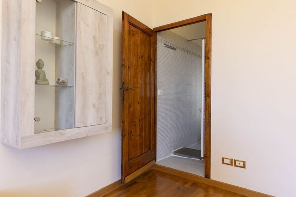 A room with a wooden door leading to a shower area, a wooden cabinet with glass shelves, and a small statue. - Deluxe Saint-Esprit Apartment (Firenze)