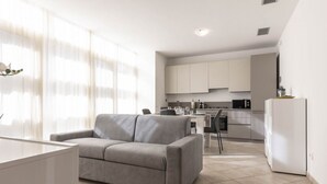 A modern living room with a grey sofa, a dining table, and a kitchen area with white cabinets and appliances.