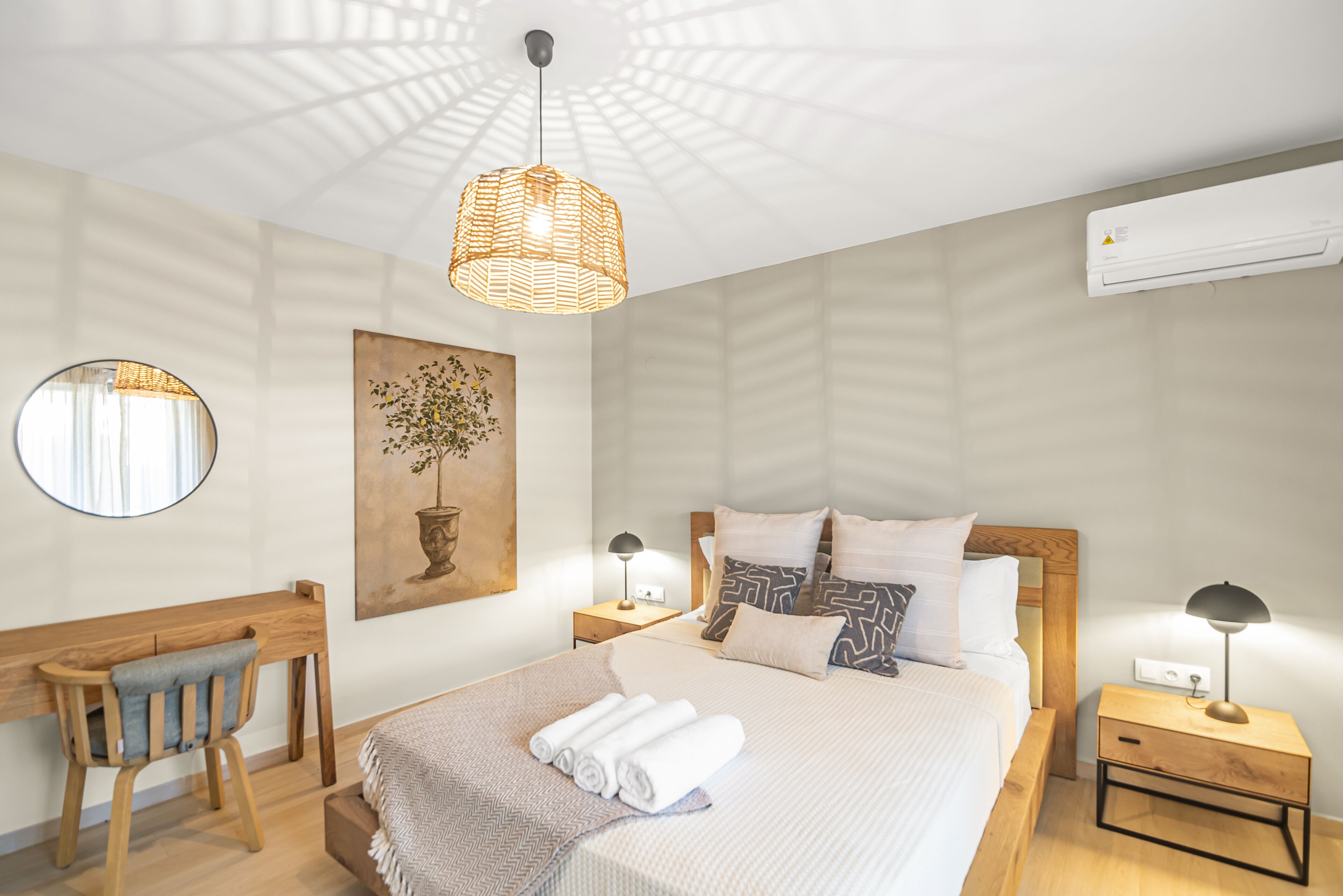 A neatly arranged hotel room with a bed, bedside tables, a desk, a chair, a hanging light fixture, and a wall-mounted air conditioner.