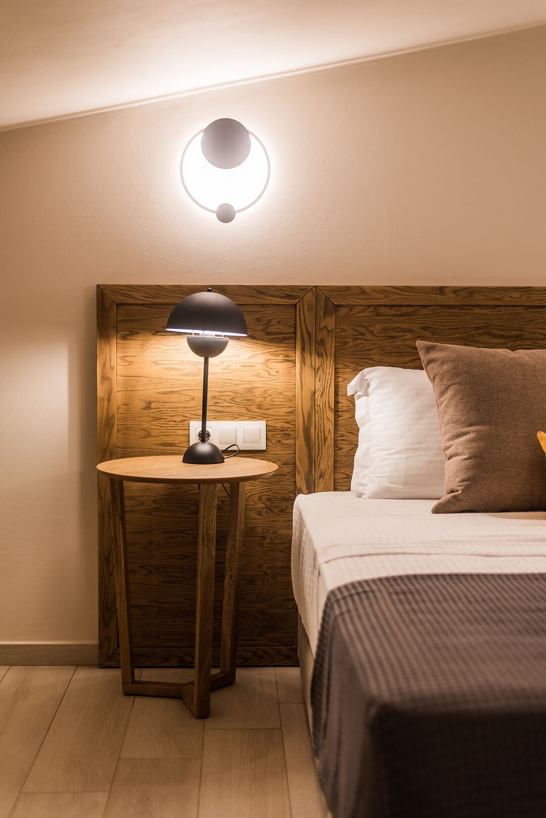 A hotel room with a wooden headboard, a round wooden nightstand with a lamp, a bed with two pillows, and a wall-mounted light fixture.