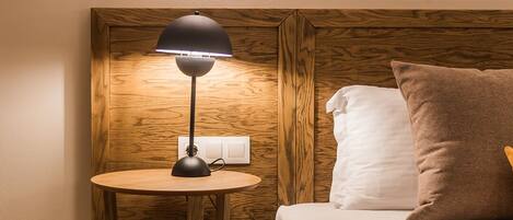 A hotel room with a wooden headboard, a round wooden nightstand with a lamp, a bed with two pillows, and a wall-mounted light fixture.