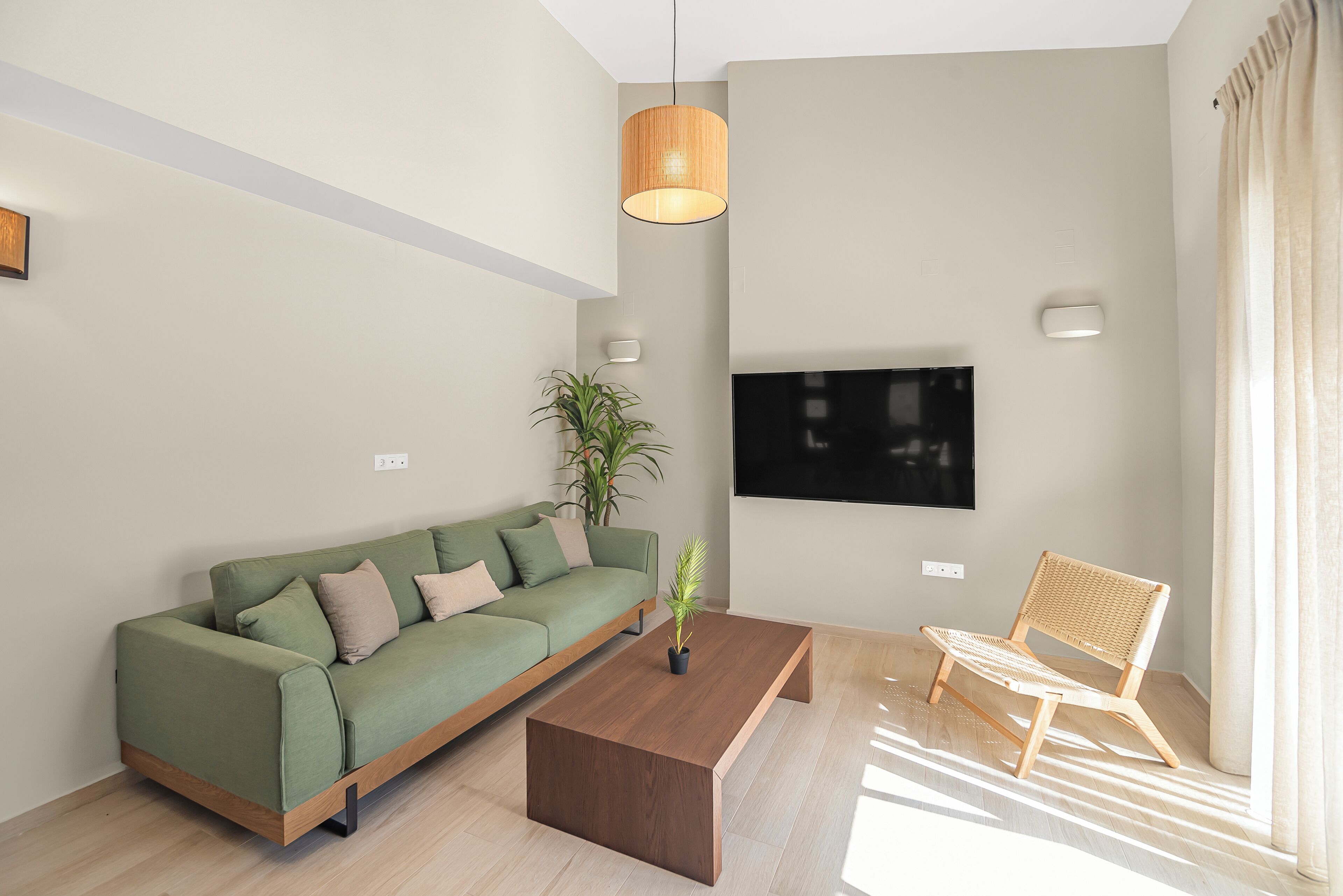 Ein modernes Wohnzimmer mit einem grünen Sofa, einem Holztisch, einem Korbstuhl, einer Topfpflanze und einem an der Wand befestigten Flachbildfernseher.
