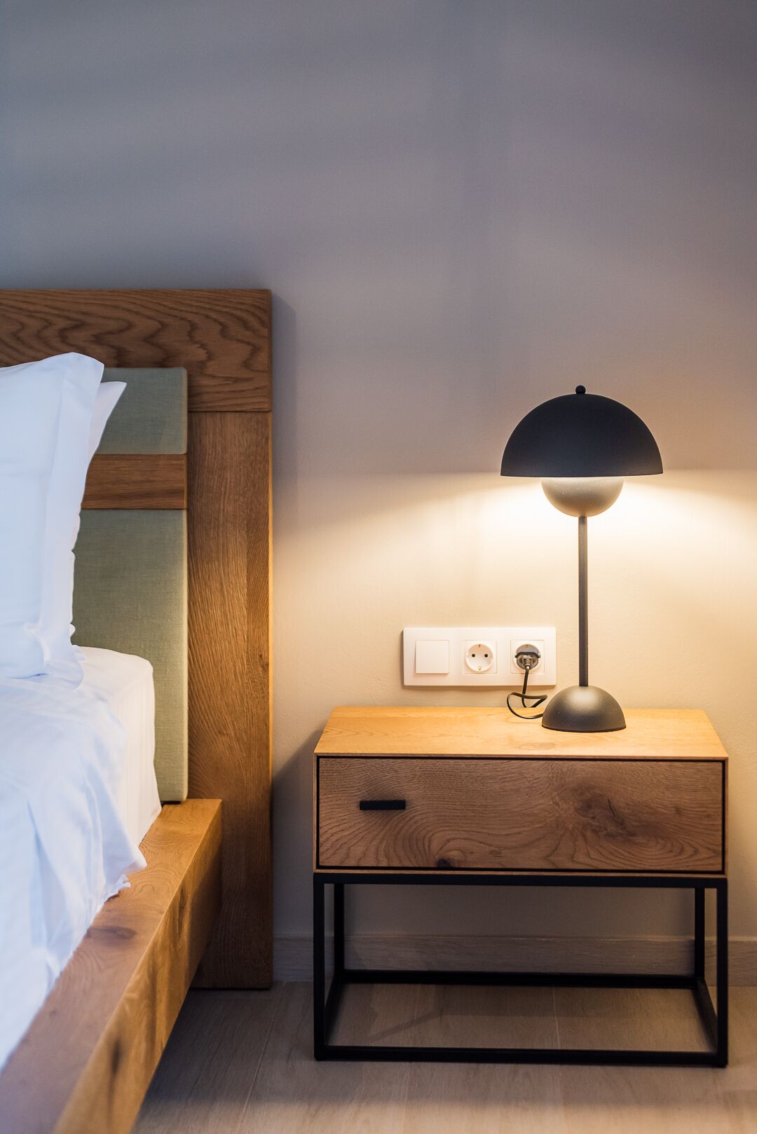 A neatly made bed with white linens, a bedside table with a lamp, and a power outlet on the wall.