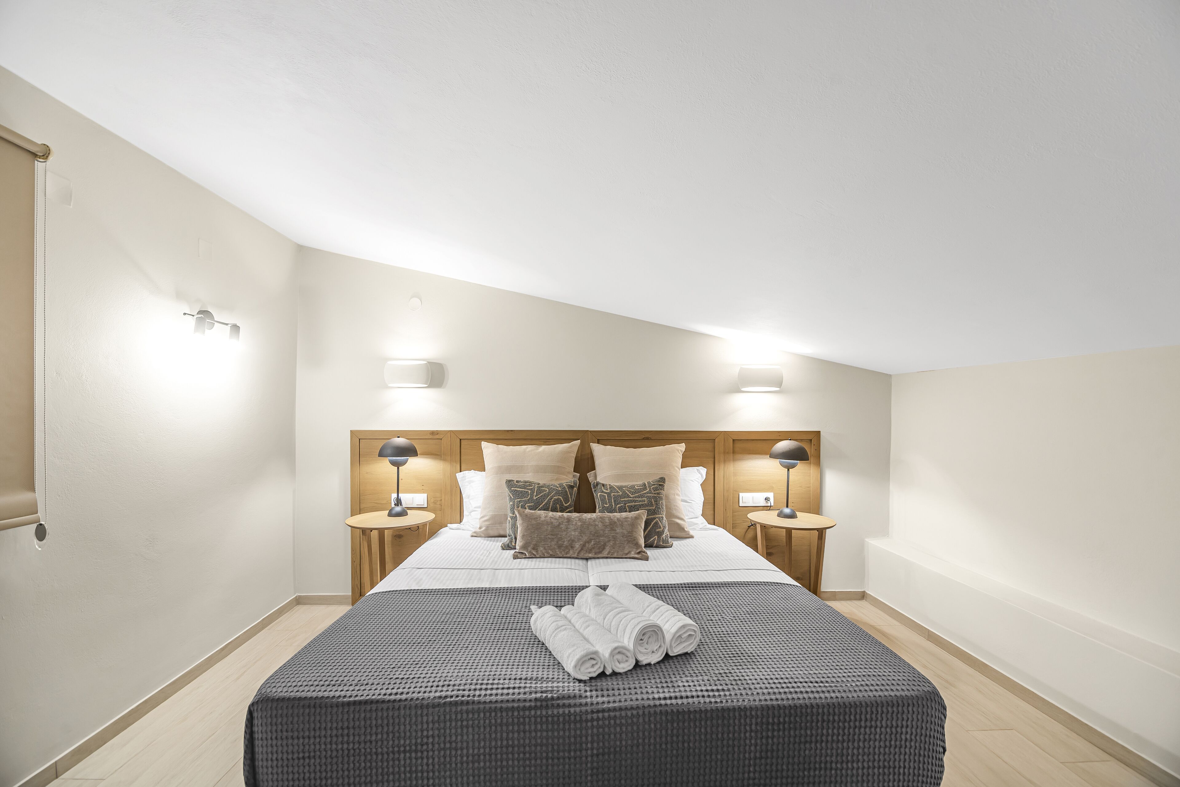 A neatly made bed with a gray bedspread and rolled towels, two bedside tables with lamps, and a wooden headboard.