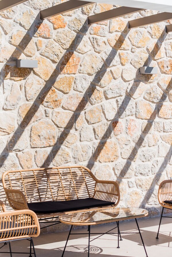 A stone wall with a geometric pattern, a wicker chair with a black cushion, and a small table. - Stone and Sea Villas (Lefkada)