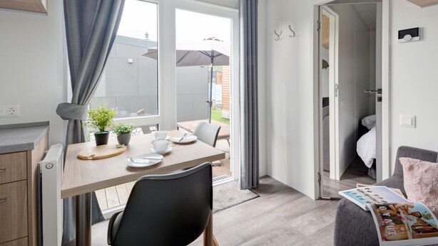 A modern, well-lit room with a wooden dining table, a black chair, and a small potted plant. There is a window with curtains, a door leading to another room, and a magazine on a cushion. - Tiny Haus Central by Interhome (Beilngries)
