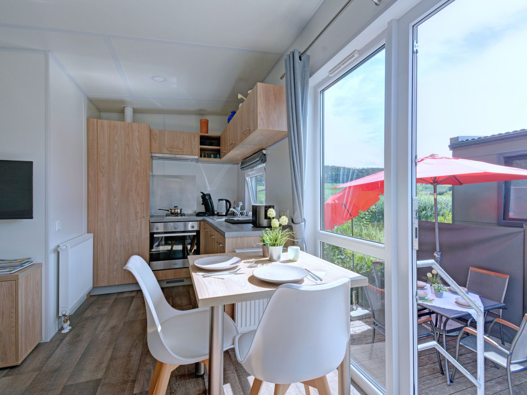A compact kitchen with wooden cabinets, a dining table with chairs, and a large window overlooking a garden.