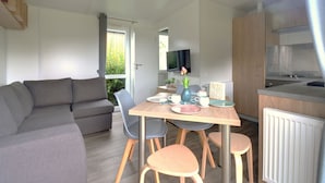 Ein modernes, kompaktes Wohnzimmer mit grauem Sofa, einem Holzesstisch mit vier Stühlen, einer kleinen Küchenzeile mit Herd und einem an der Wand befestigten Fernseher.