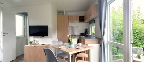 A modern kitchen with wooden cabinets, a dining area with a table and chairs, a television mounted on the wall, and large windows offering a view of greenery.