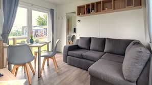 Ein modernes Wohnzimmer mit grauem Sofa, Esstisch aus Holz und Stühlen. Ein Fenster mit Vorhängen, ein Bücherregal mit Büchern und Pflanzen, sowie ein an der Wand befestigter Fernseher.