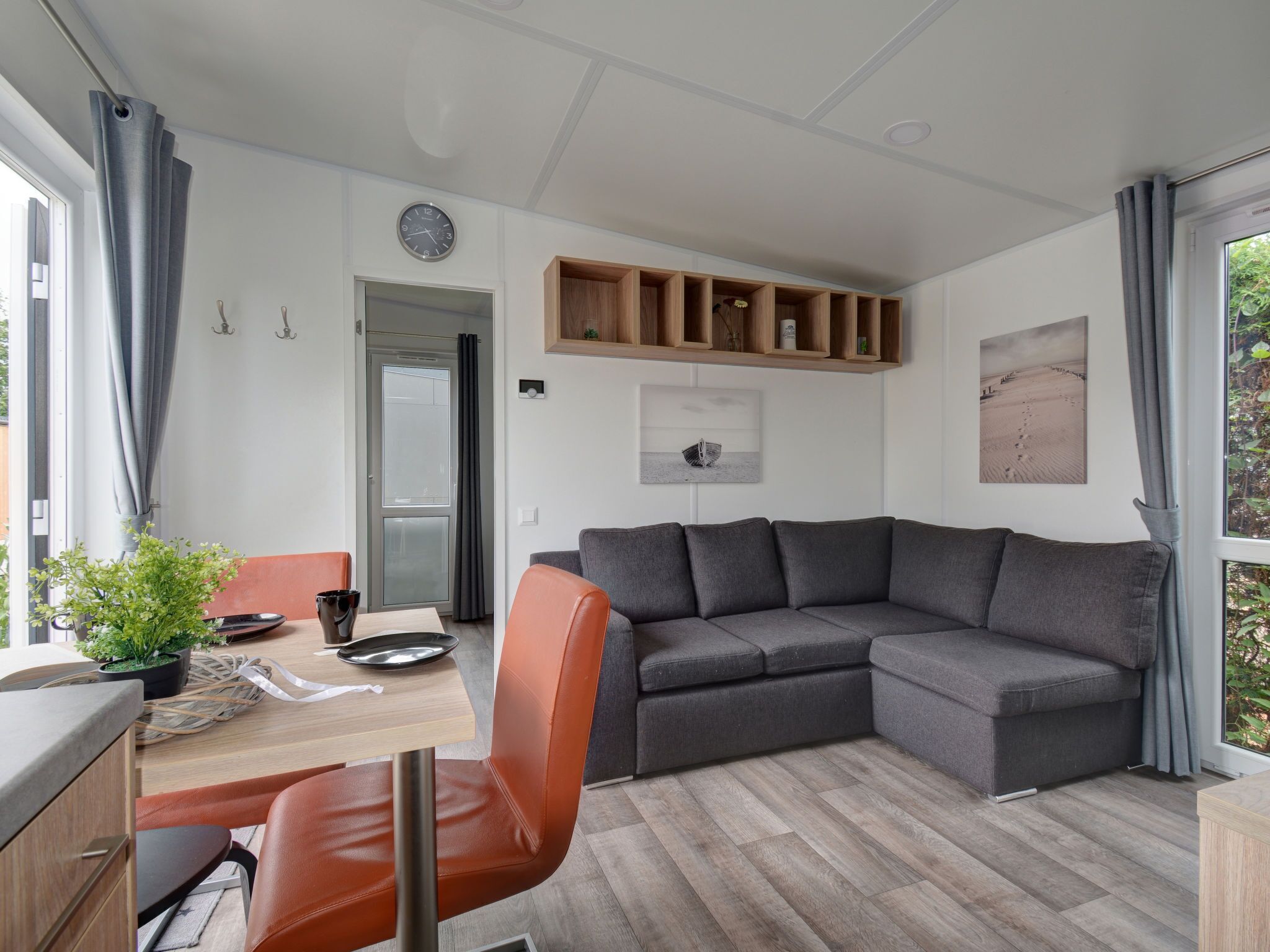 A modern living room with a grey sectional sofa, a wooden dining table with chairs, a small potted plant, and a wall-mounted clock.
