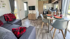 Een moderne woonkamer met een grijze bank en rode geruite kussens, een eethoek met een houten tafel en stoelen, en een keuken met houten kastjes en een inbouwoven.