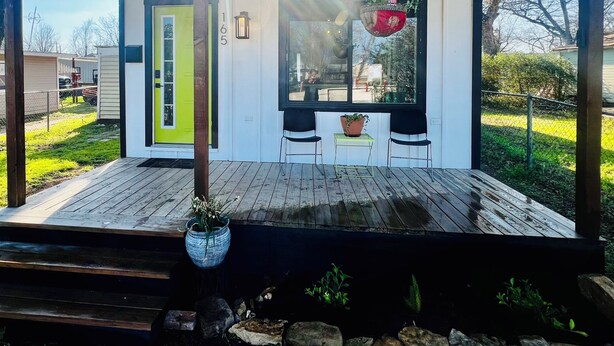 Un espace extérieur abrité, doté d’une terrasse en bois, d’une porte verte et d’une fenêtre ornée d’une plante suspendue.