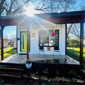 A covered outdoor area with a wooden deck, a green door, and a window with a hanging plant.