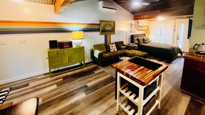 A cozy room with a bed, a green sofa, a wooden table with a kettle, and a small kitchen area.