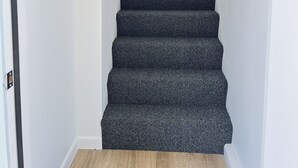 Un escalier avec des marches recouvertes d’une moquette sombre et une rampe noire, menant à un palier orné d’un tapis aux motifs bleus et blancs.