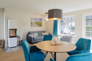 Una sala de estar moderna con un sofá gris, sillas azules, una mesa redonda de madera y un ventanal con vistas al exterior.