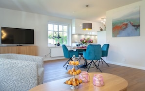 Una sala de estar moderna con televisor de pantalla plana, un sofá, un comedor con sillas azules y una cocina al fondo.