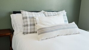 Cama com roupa de cama branca e duas almofadas, uma estampada xadrez e outra listrada, em frente a uma parede verde.