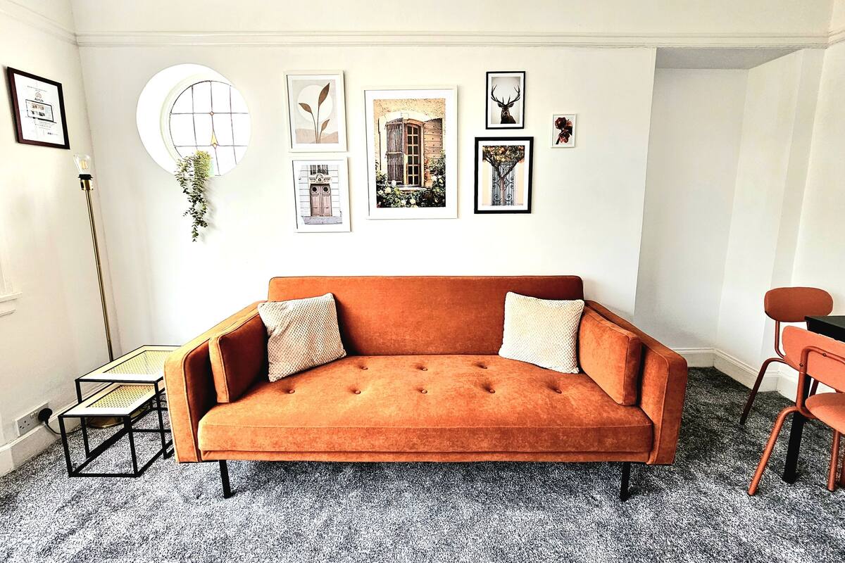 A room with a brown sofa, a small table, and a chair. There are framed pictures on the wall.