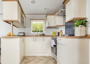 Una cocina compacta con armarios blancos, piso con motivo de espiga, ventana con persianas y una planta sobre la encimera.