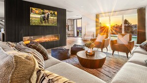 A modern living room with a fireplace, comfortable seating, and a large window with a sunset view. - Spacious Retreat with Heated Pool-Hot Tub-Games-Bar-Near Keeneland&Bourbon Trail (Lexington)