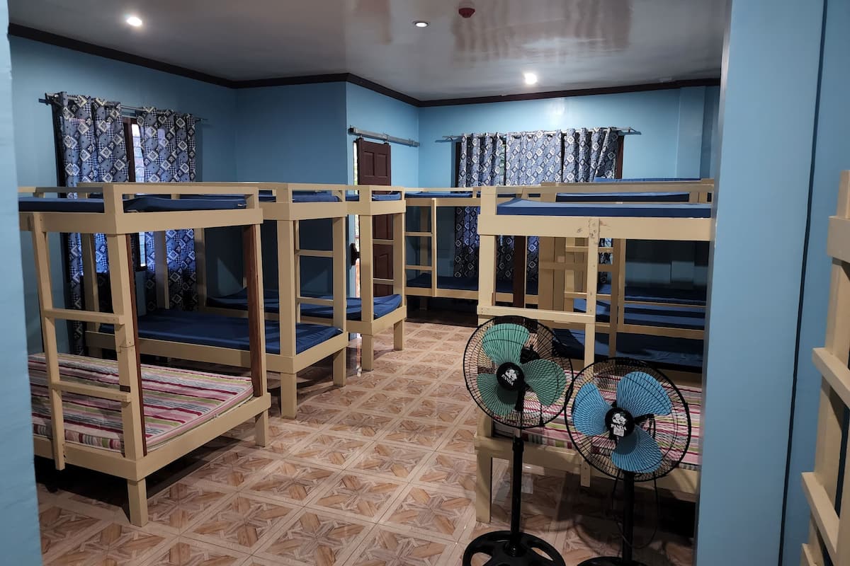 Ein Schlafsaalzimmer mit Etagenbetten, gemusterten Vorhängen und Standventilatoren.