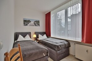 Quarto de hotel com duas camas, janela com cortinas leves, um quadro na parede e um radiador.