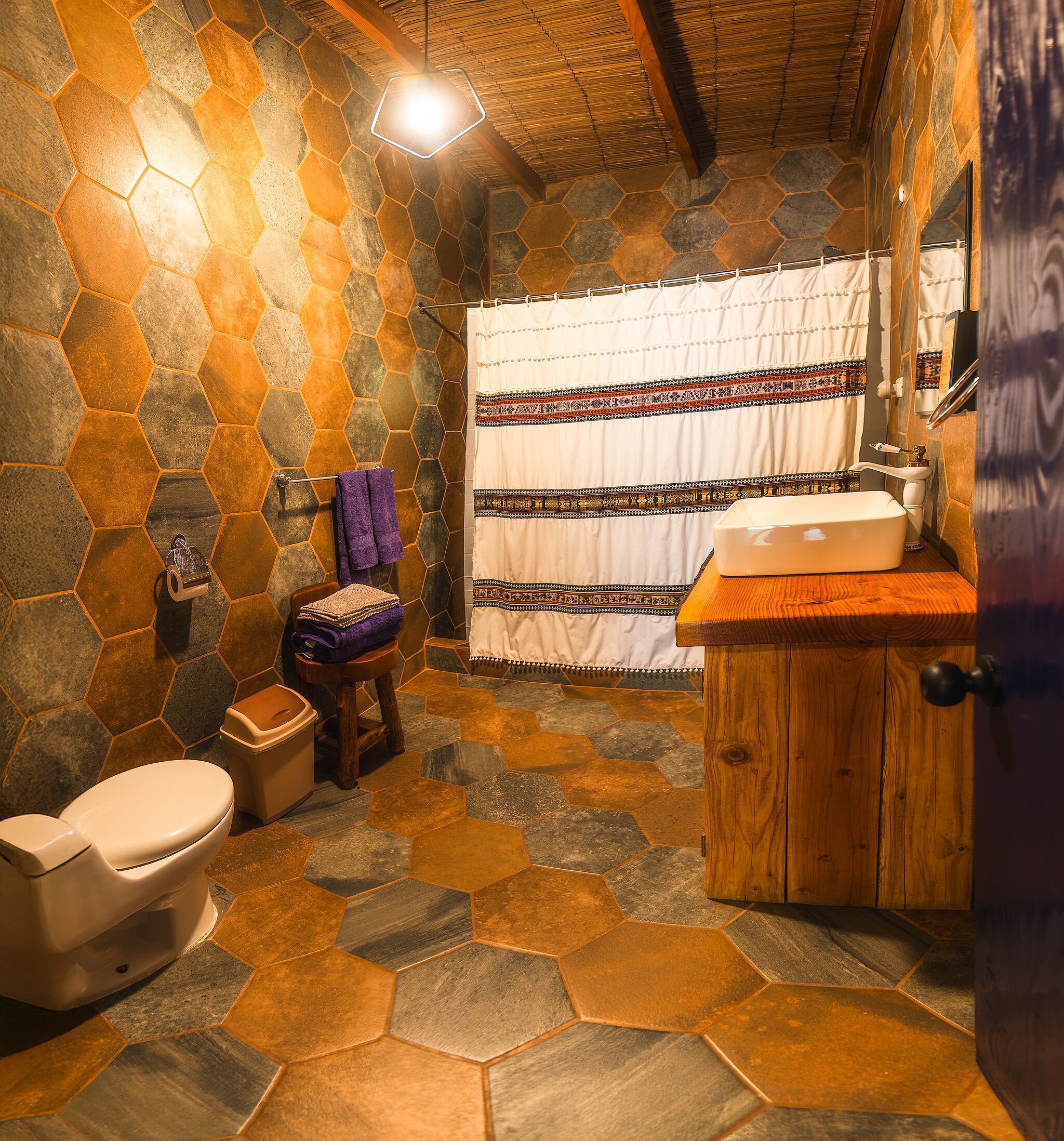 A bathroom with hexagonal stone tiles, a wooden vanity, a toilet, and a shower curtain with a patterned border.