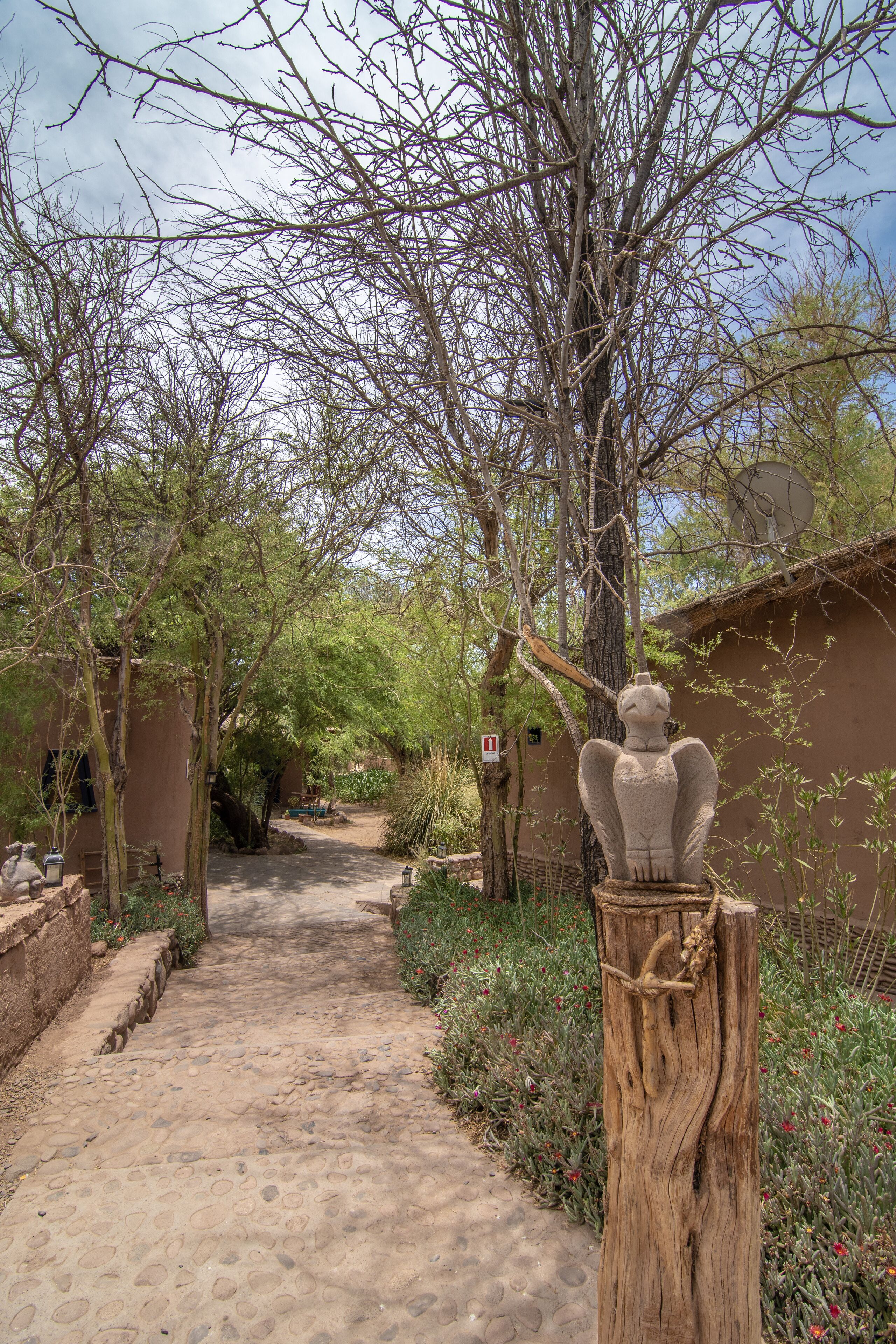 A cobblestone path leading through a garden with a wooden sculpture and a building in the background.