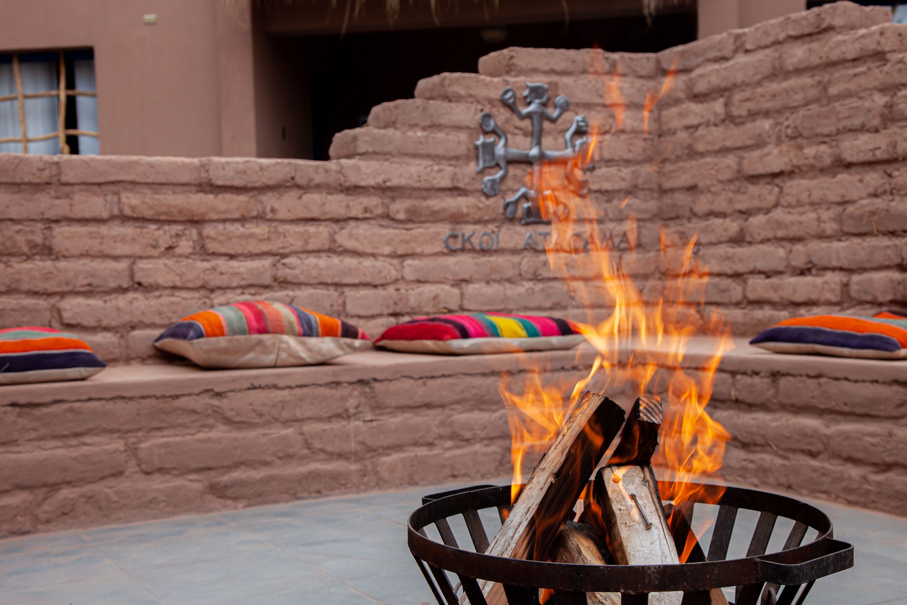 Un foyer avec des bûches et des flammes, un mur de briques et des coussins colorés sur un banc.
