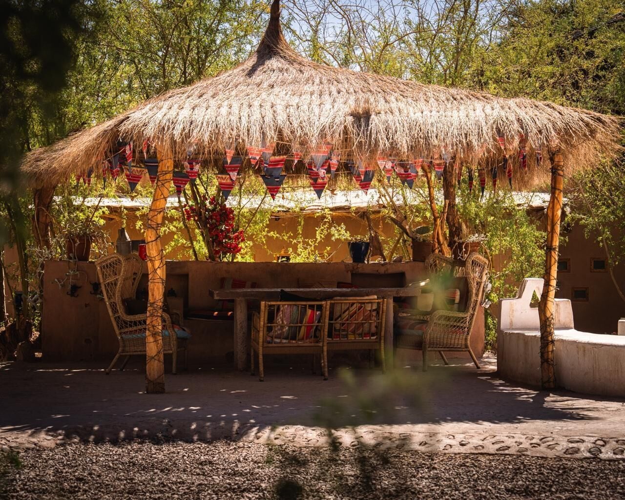 Un espace extérieur aménagé avec des sièges, doté d'un toit de chaume, de chaises en osier et d'une table, le tout entouré d'arbres et bordé d'un sol sablonneux.