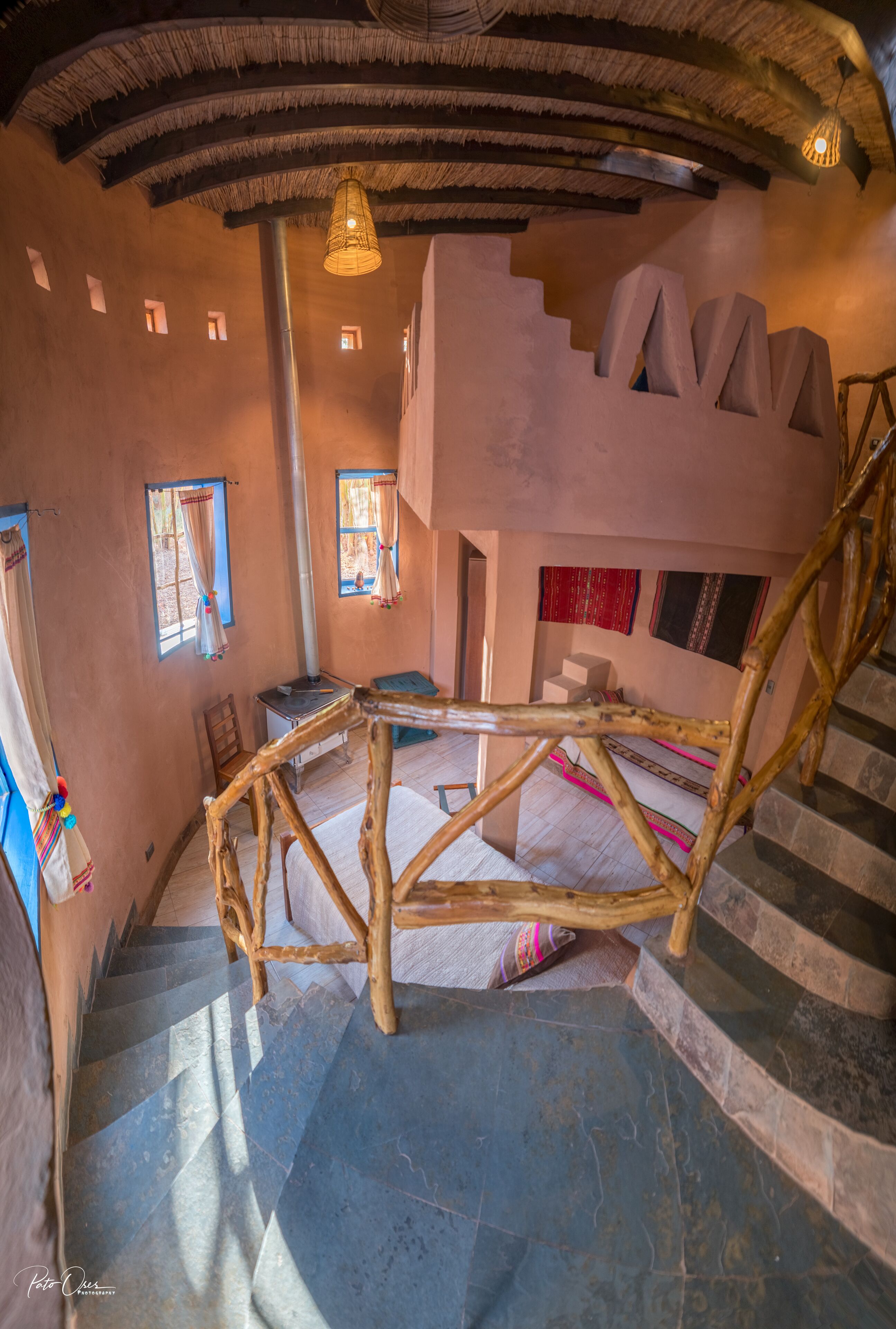 A rustic interior with a wooden staircase, a thatched ceiling, and a stone wall with a unique geometric design.