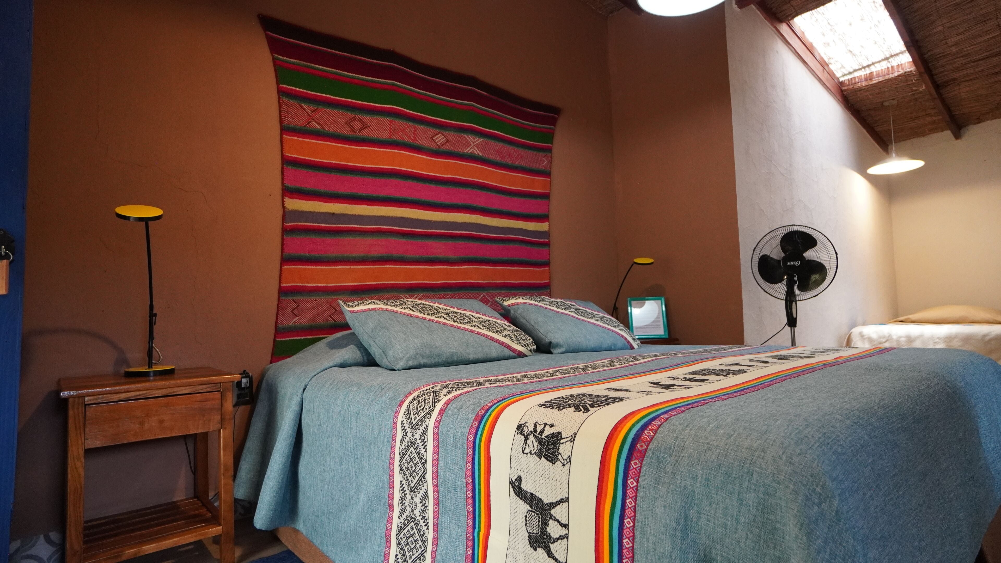 Un dormitorio con cama, mesita de noche, ventilador de pared y un tapiz colorido.