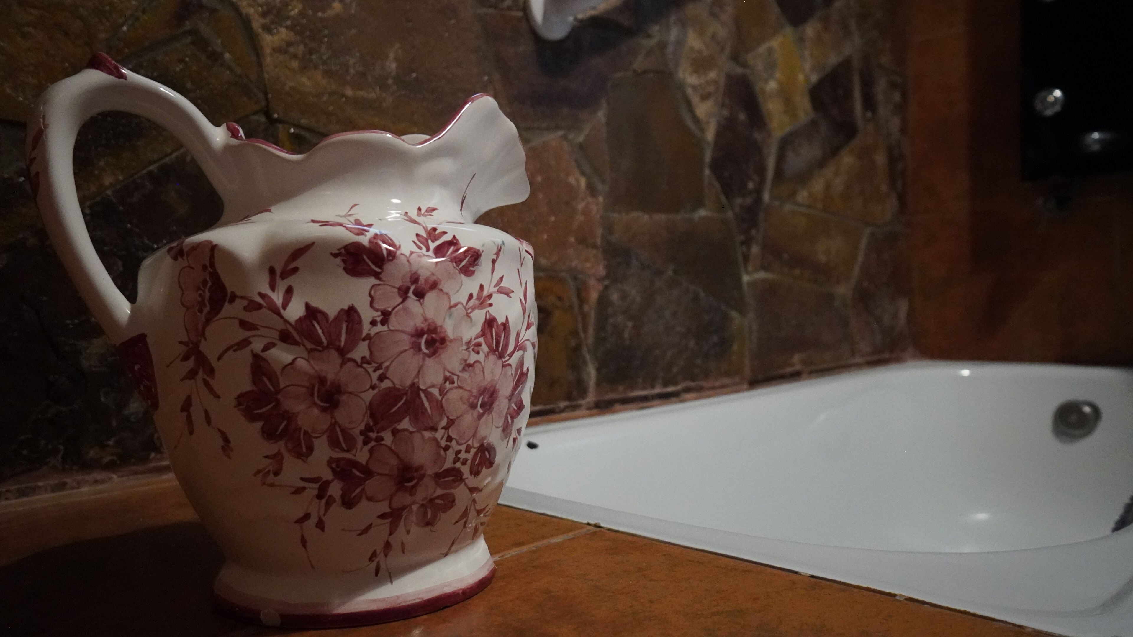 A white ceramic pitcher with floral designs on a countertop next to a bathtub.