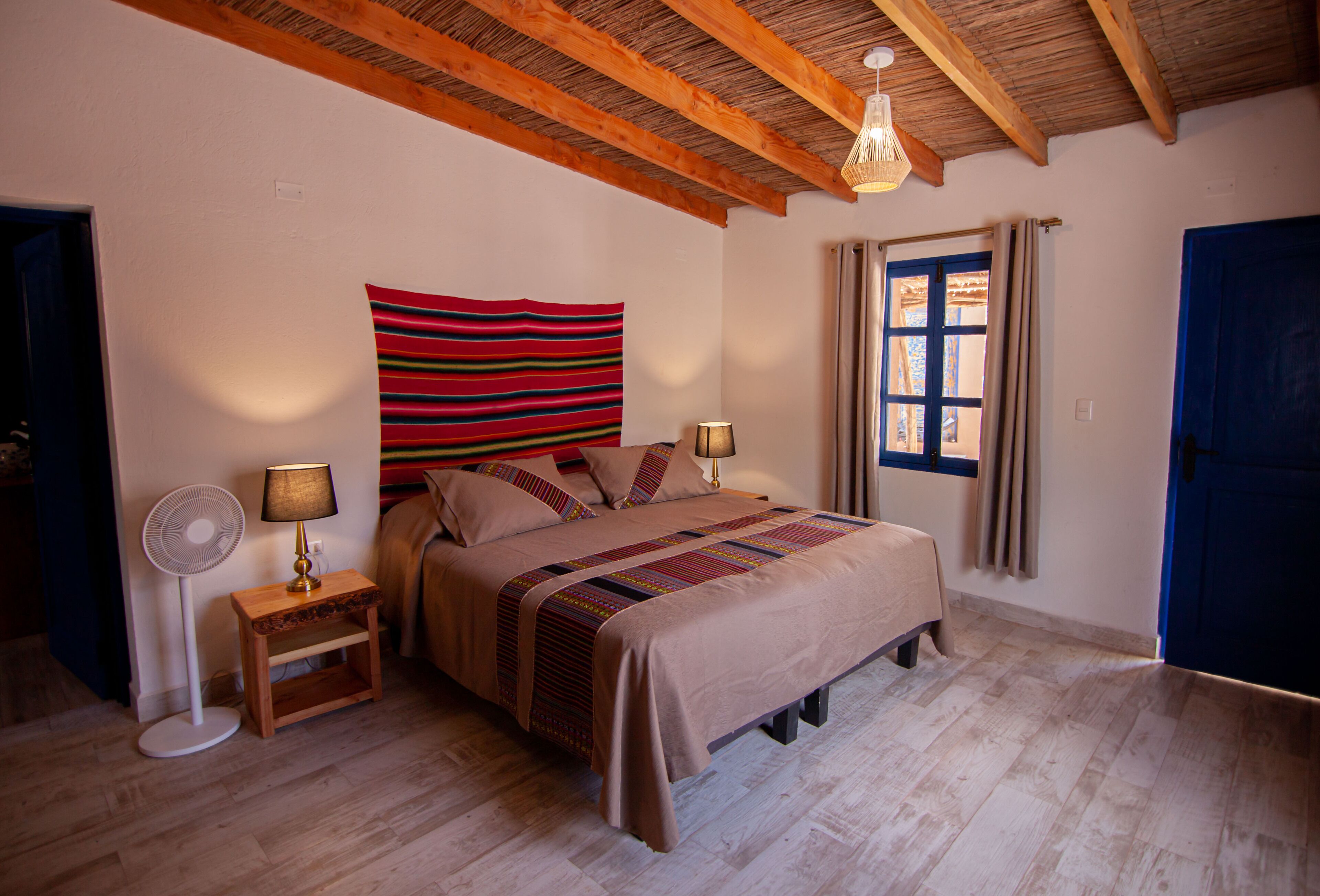 Un dormitorio con cama, lámparas de noche, un ventilador, una ventana con cortinas y una puerta azul.
