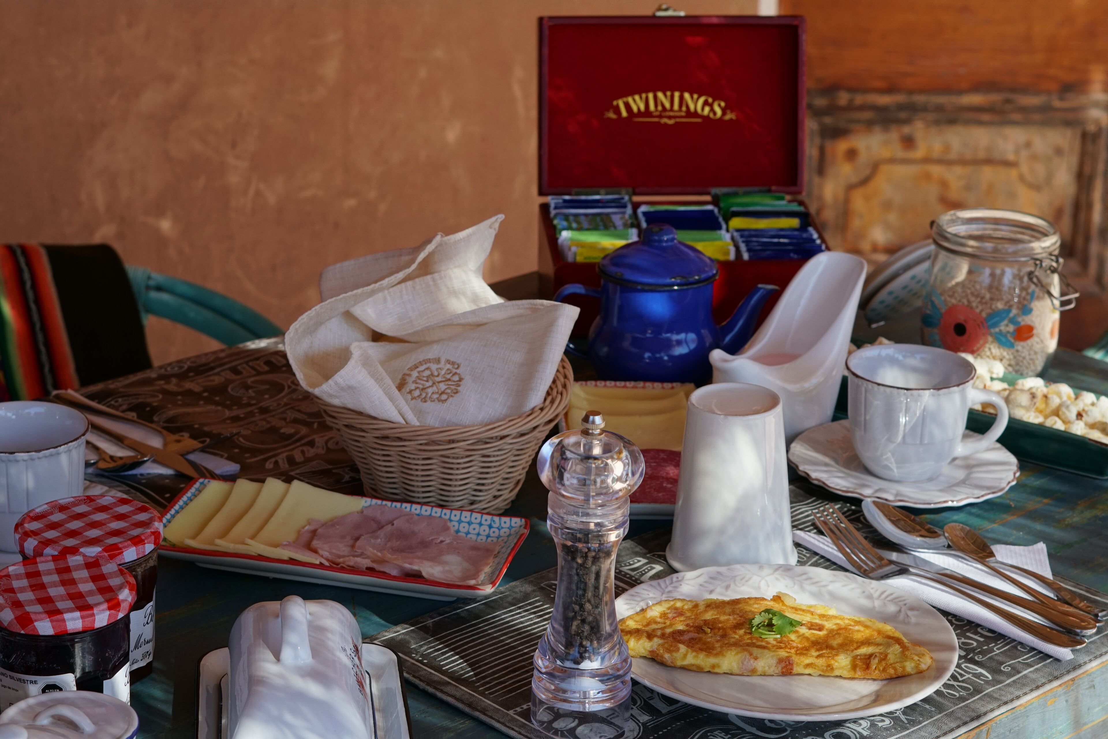 Una mesa de desayuno con un plato de jamón, queso y una tortilla, junto con condimentos, una cafetera y una caja de té Twinings.