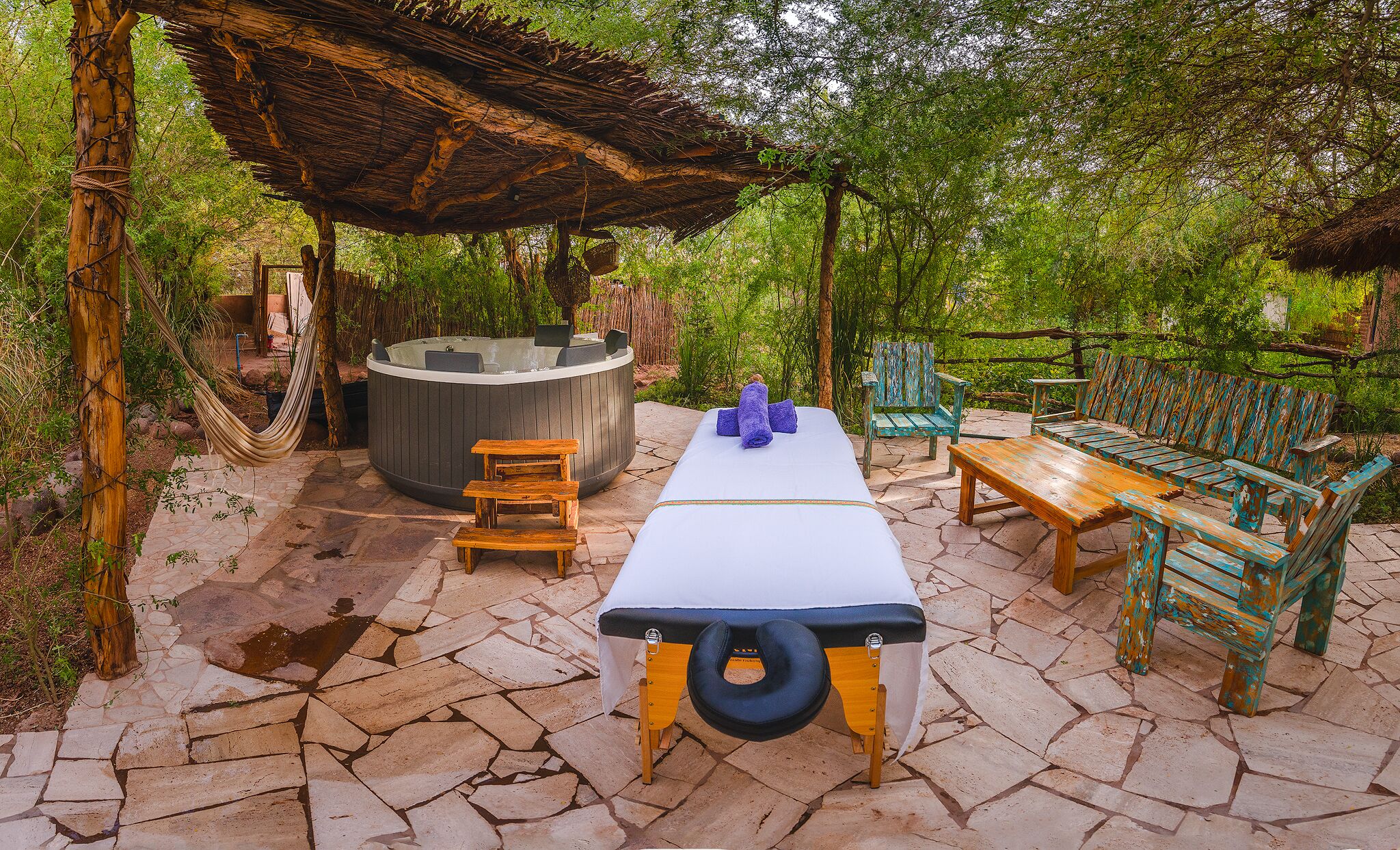 An outdoor relaxation area with a hot tub, wooden seating, and a stone-tiled floor.