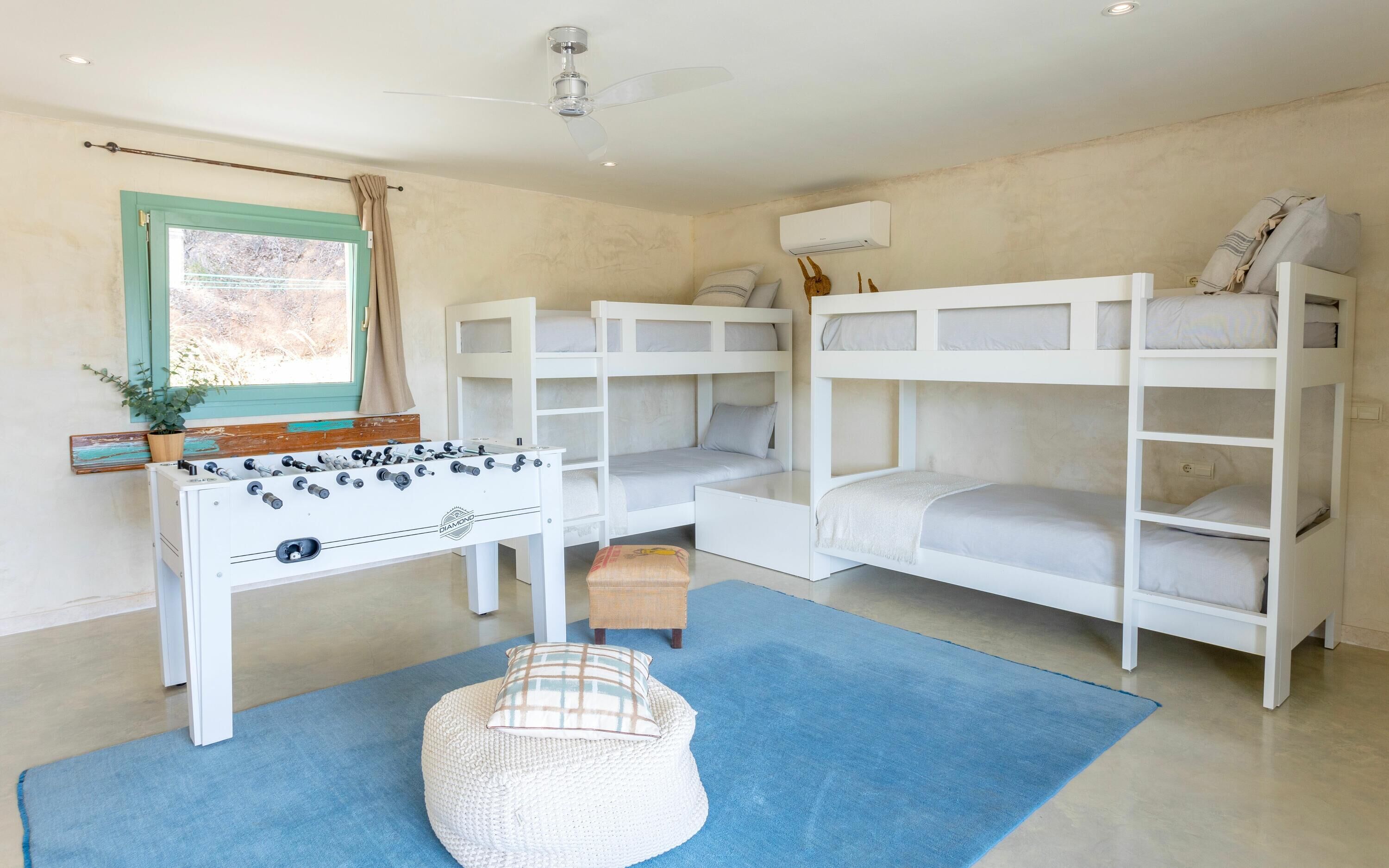 A room with bunk beds, a foosball table, a window with curtains, and a ceiling fan.