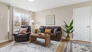 A living room with a leather sofa, a recliner, a coffee table, a potted plant, and a large window with curtains. - NEW! Cozy 1BR Flat w/ King Bed & Workspace (Roseville)