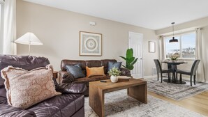 A living room with a brown leather sofa, a wooden coffee table, a large potted plant, and a dining area with a table and chairs. - NEW! Cozy 1BR Flat w/ King Bed & Workspace (Roseville)
