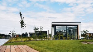 Un bâtiment moderne doté de grandes fenêtres en verre, d’une terrasse en bois et d’un espace verdoyant.