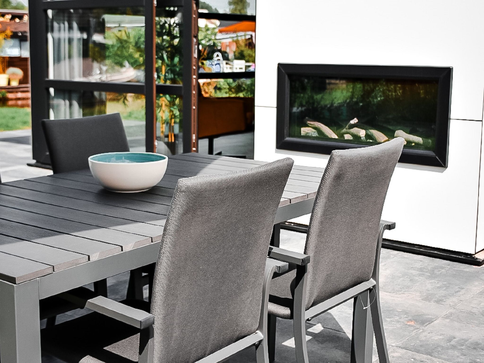 A modern outdoor dining area with a black table and grey chairs, a white bowl on the table, and a large window with a view of greenery.
