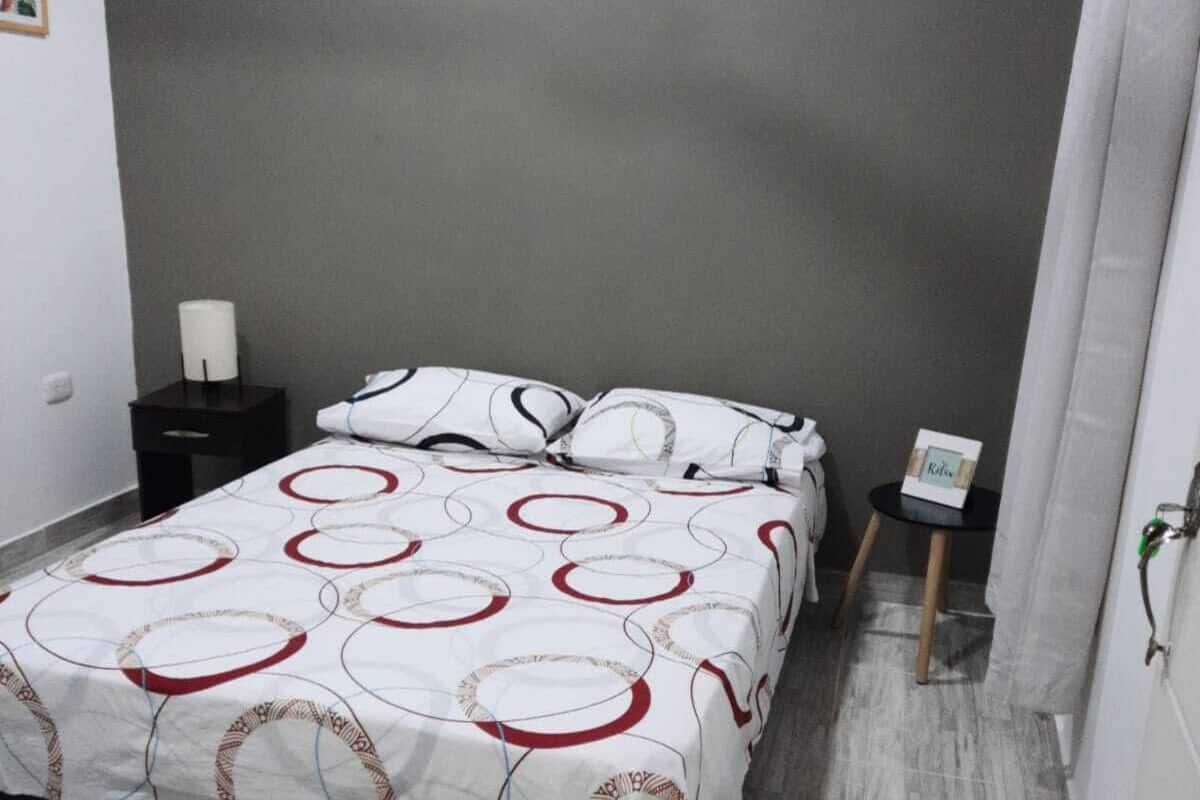 A bedroom with a bed, a nightstand, a small table, and a wall-mounted light.