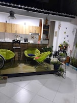 Una cocina con armarios de madera, una silla negra y una silla verde con cojín. Hay plantas en macetas y una pequeña mesa con una silla.