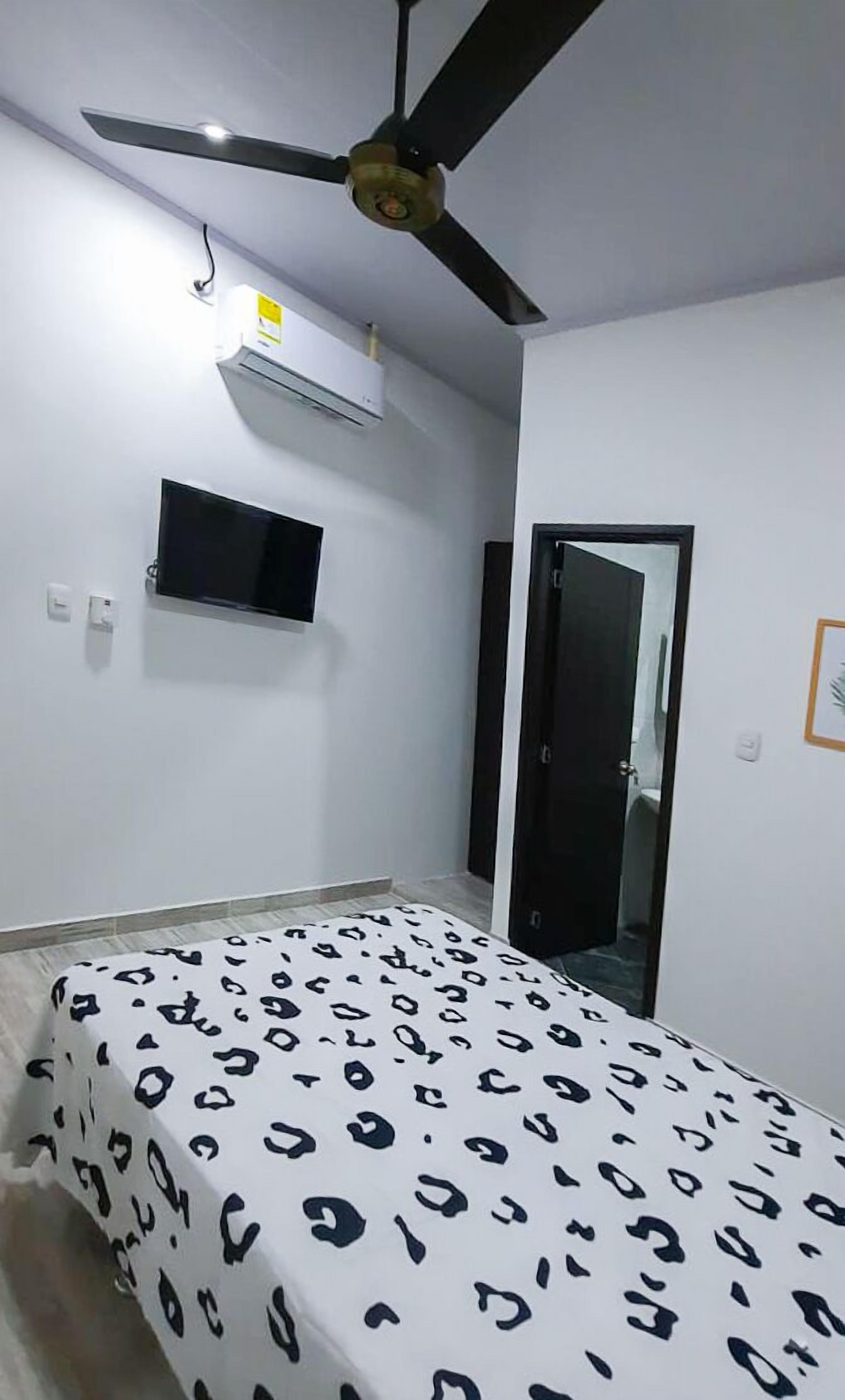Ein Schlafzimmer mit Bett, Deckenventilator, Klimaanlage, einem an der Wand befestigten Fernseher und einer Tür.