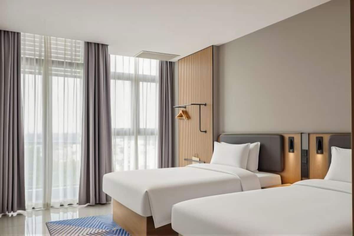 Une chambre d’hôtel avec deux lits, une grande fenêtre avec des rideaux, un lampadaire fixé au mur et un décor mural en panneaux de bois.